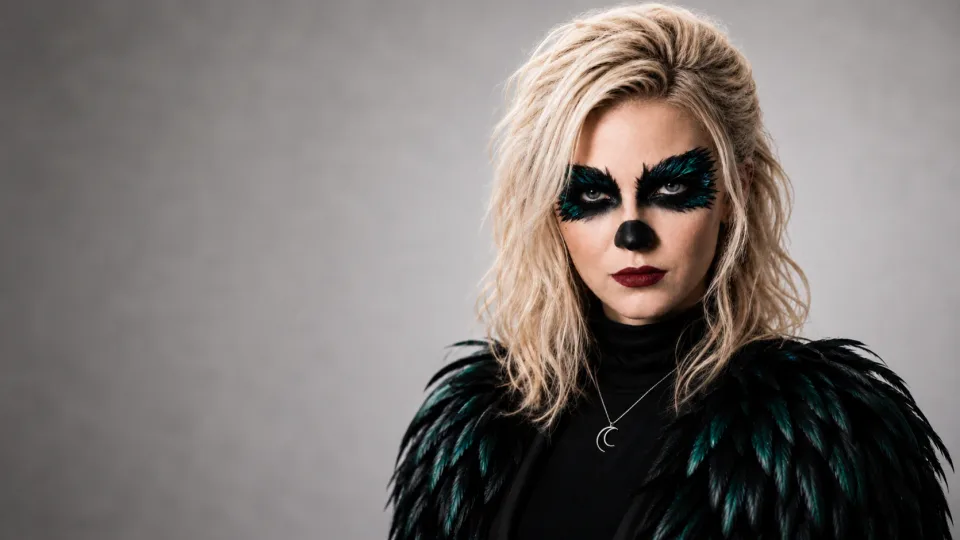 Clarapetra in a black feathered costume and teal raven-inspired makeup against a pale studio background.